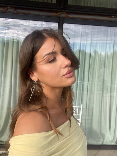 A woman with brown hair and fair skin is looking up at the camera. She is wearing a light yellow dress and a silver earring. The background is a window with a view of trees and sky. This image was taken in the city of Беране, Montenegro.