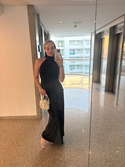 A woman stands in a hallway, taking a mirror selfie. She is wearing a black sleeveless dress, a pearl headpiece, and clear mesh shoes. She holds a white pearl-beaded handbag in her left hand and her phone in her right. The hallway has a speckled granite floor and a wall with recessed lighting. A large window to her left reflects a white building with many windows. The scene appears to be indoors, possibly in a hotel or an apartment building. The lighting suggests it is daytime. There is no visible text in the image.