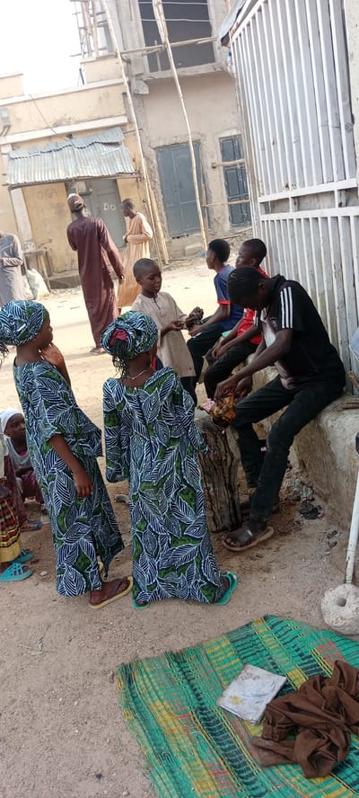 The image depicts a group of individuals in Kwachiri, Nigeria, gathered in an outdoor setting. Several children stand to the left, two of whom are wearing identical dresses with a leafy blue, green, and white pattern. In the center, a boy is holding a piece of meat. To the right, a young man wearing a black shirt with white stripes on the sleeves and black jeans is sitting on a concrete structure, cutting up meat on a wooden stump. Behind him, two other boys are seated. In the background, there are buildings, one with scaffolding. In the foreground, there is a patterned mat on the ground with a book and brown fabric on top of it. There are also other individuals in the background, some wearing traditional Nigerian clothing.