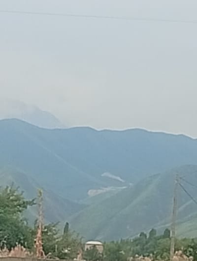 The image shows a hazy, mountainous landscape with rolling green hills. The sky is overcast and pale blue. In the foreground, there is a row of trees and shrubs, with a faint outline of buildings visible through them. A tall, thin utility pole with wires stands to the right of the frame. The scene appears to be in a rural or semi-rural area, possibly the outskirts of Besh Kungei, Kyrgyzstan. There are no people or animals visible in the image. The overall impression is one of a quiet, natural setting.
