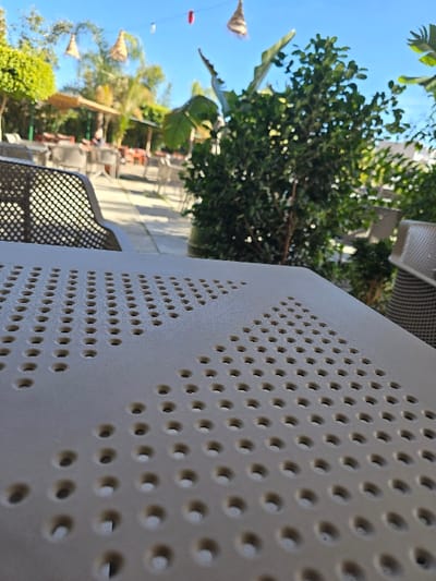 This image depicts an outdoor seating area of an establishment in Taza, Morocco, under clear, bright daylight. The foreground is dominated by a beige, matte table surface, textured with numerous circular perforations arranged in a grid pattern, some areas forming distinct triangular or chevron designs. In the midground, blurred dark grey or black grid-patterned chairs and tables are visible, indicating an organized seating arrangement. Lush green foliage, including a prominent bush on the right, partially obscures the view of the background. The background features a vibrant blue sky overhead, adorned with decorative elements such as several conical, straw-colored lampshades, a single red spherical lantern, and string lights suspended across the outdoor space. Additional blurry trees, possibly with palm-like fronds, and canopy structures providing shade are also present. No individuals are clearly identifiable; extremely blurred figures may be present among the tables in the distant background. The scene is illuminated by strong natural sunlight.