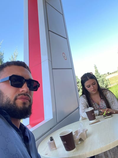 Two individuals are seated at a circular table outdoors. In the foreground, a man wearing black sunglasses and a dark blue patterned shirt is in profile, looking to his left. To his right, a woman with dark hair, wearing a white and gold embroidered top, looks down at her phone. Two disposable coffee cups and a vaping device are on the table. Behind them, a large, modern structure with red and white panels stands against a clear blue sky. The surrounding area appears to be a park or green space with trees and grass. The scene is likely located in the city of Korkmaskala, Russia, as suggested by the image context.