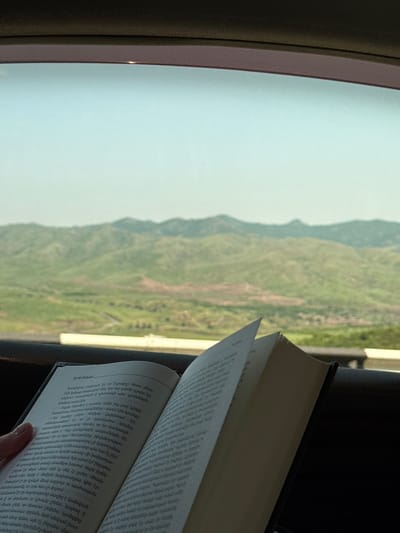 A person's left hand is holding an open book, with text in Armenian, positioned in the foreground. The book's pages are slightly fanned out. Behind the book, a blurred landscape of green, rolling hills and distant mountains under a pale blue sky is visible. The scene appears to be captured from inside a vehicle, suggested by the dark interior framing the view. The location is identified as Voskepar, Armenia.