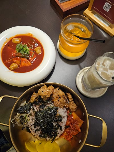 A high-angle, overhead shot shows a table with several Korean dishes and drinks. In the foreground, a brass-colored bowl contains rice topped with dried seaweed, bean sprouts, fried chicken, and kimchi. To the left, a white bowl holds a red, stew-like dish with fish cakes and green onions. Two glasses are visible: one filled with a golden-colored beverage with ice and a straw, and another with a milky white drink and ice. A wooden stand with a QR code is in the background. The setting appears to be a restaurant, potentially in Ngaglik, Indonesia.