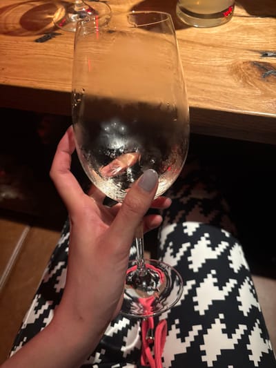 The image displays a close-up, high-angle perspective of an individual's right hand holding a clear, stemmed wine glass. The hand has fair skin, and the fingernails are manicured and painted a light grey color. The glass contains a clear, colorless liquid, approximately half-full, exhibiting external condensation and numerous small bubbles within. A distinct plume of white mist or smoke is visible rising from the liquid inside the glass, consistent with the use of dry ice.
The individual is wearing garments with a black and white geometric, pixelated pattern, visible across the lap and thighs. A bright pink cord or strap is partially visible in the lower center foreground, below the glass. The glass is positioned over a rustic wooden table surface, characterized by visible grain, knots, and dark striations. Reflections of a red light source are present on the table and on the base of an inverted, empty glass stem partially visible in the upper left background. Another blurry, cylindrical glass containing a pale yellow liquid is partially visible on the table in the upper right. The scene is set indoors, likely within a restaurant or bar environment, in Sankt Anton am Arlberg, Austria.