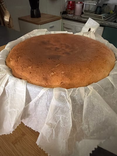 A top-down view depicts a freshly baked loaf of bread, golden-brown in color with a slightly uneven surface texture, nestled within a white parchment paper lining. The bread rests on a wooden surface, likely a countertop or cutting board. To the left of the bread, part of a white cabinet and a black object (possibly a shoe or kitchen utensil) are visible. Above the cabinet, a section of a metal rack containing assorted items, including a red beverage can, is discernible. The environment suggests a kitchen setting. There is no indication of the image having been taken in Azuqueca de Henares, Spain.