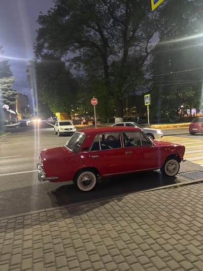 A red vintage car is parked on the side of a street next to a cobblestone sidewalk. The location is indicated by a pedestrian crossing sign visible nearby. The background features trees and buildings, with streetlights illuminating the evening scene. A white vehicle is parked in front of the red one. The sign "Komgaron" is visible in the distance suggesting the location within the city of Komgaron, Russia.