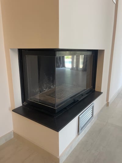 A modern corner fireplace with a glass front is situated in a room with beige and white walls and a tiled floor. The fireplace has a black metal frame and a black hearth. To the right of the fireplace, a silver vent is installed in the wall. The room appears to be a living space, possibly in a residential property. The image does not provide any specific landmarks or context to determine its location within Moni Community, Cyprus.