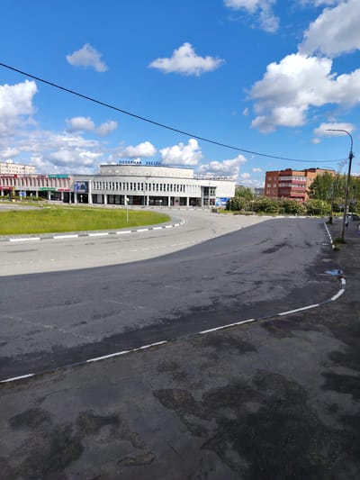 The image depicts a street scene in Olenegorsk, Russia, featuring a large building with the inscription "ПОЛЯРНАЯ ЗВЕЗДА" (Polar Star) prominently displayed across its facade. The building appears to be a modern structure with a white facade and numerous windows. In front of the building, a circular area with green grass and yellow wildflowers is visible, bordered by a paved road with white and black striped curbs. The road curves to the right, leading away from the building and into the foreground.
Several other buildings are present in the background, including a multi-story brick residential building and other commercial structures. A lamppost with a decorative reindeer figure is situated on the right side of the road, and a few individuals and a vehicle are sparsely distributed along the sidewalk. The sky is a clear blue with scattered white clouds, indicating fair weather. The overall impression is of a relatively quiet urban environment on a sunny day.