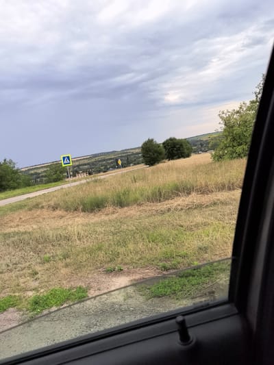 The image captures a roadside view from inside a vehicle, looking out through the open window. The scene is set in a rural or semi-rural area, likely near Belogorskiy, Russia, given the context. A paved road runs diagonally from the left to the center of the frame, bordered by dry, tall grass and vegetation. A yellow pedestrian crossing sign, featuring a blue silhouette of a person, is prominently visible on the left side of the road. Further down the road, another yellow warning sign, possibly indicating a hazard or change in road conditions, is situated. The landscape beyond the road consists of rolling hills covered with trees and fields, under a cloudy sky. The foreground of the image shows the interior of the car, including the car door and a portion of the window. The overall impression is a snapshot of a journey through a natural landscape under a somber sky.