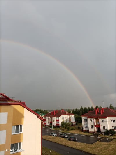 A wide-angle, vertical outdoor shot captures a post-rain scene in the village of Ulukavetsky Selsoviet, Belarus. The sky is overcast and a double rainbow is visible, arching across the upper half of the frame. In the foreground, on the left, is the side of a multi-story residential building, painted in alternating bands of pale yellow and white, with visible windows. The building has a red-tiled roof. To the right of this building are two more residential buildings, also with red-tiled roofs, appearing to be apartment complexes with multiple floors and windows. They are constructed with white brickwork and accents of yellow and red. A wet asphalt road curves in front of the buildings, reflecting the grey sky. Several cars are parked on the side of the road or on designated parking areas. A line of trees forms a backdrop behind the buildings, with a few more trees interspersed in the yards. The ground in front of the buildings is covered in grass and some patches of mud, indicating recent rain. The scene appears to be a residential neighborhood. The bottom left corner of the image displays the text "A1 Alpha 20+".