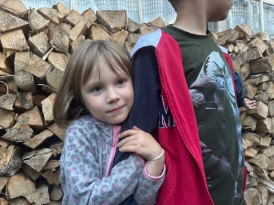 This media file shows two young children standing in front of a large stack of cut logs. The child on the left is a girl with blonde hair, wearing a grey and pink patterned fleece jacket. She is looking towards the camera with a gentle expression. Her right hand is resting on the arm of the child next to her. The child on the right is partially visible and appears to be a boy. He is wearing a dark green t-shirt with a large graphic of an eagle's head on it, and a red hooded jacket over it. The logs are stacked unevenly and appear to be of various sizes, suggesting they are prepared for firewood. The background shows a light-colored textured curtain or screen. The scene appears to be outdoors, possibly in a rural setting, given the presence of the firewood. There is no text visible in the image. The lighting suggests it could be daytime.