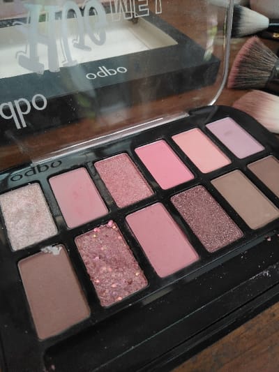 An open black odbo-branded eyeshadow palette with a clear, partially raised lid is centered on a dark, possibly wooden, surface. The palette features twelve rectangular pans in various shades of pink, brown, and glittery tones. Visible colors include: a shimmery light pink, matte light pink, shimmery medium pink, matte medium pink, matte pale peach, matte light lavender, matte light brown with a small white residue, a textured shimmery dark pink with visible glitter, matte medium pink, shimmery deep brown, and matte taupe. Scattered loose glitter and powder particles are present on the palette's black casing and the surrounding surface. Behind the palette's lid, a separate black compact, also bearing the "odbo" brand and containing white powder, is partially visible. To the right, several makeup brushes with light-colored bristles are lying on the surface. No individuals are present, and no active interactions are depicted. The scene is consistent with an indoor setting, likely a vanity or dressing table. The specified location of Шөптікөл ауылдық округі, Kazakhstan, cannot be visually verified from the image content.
