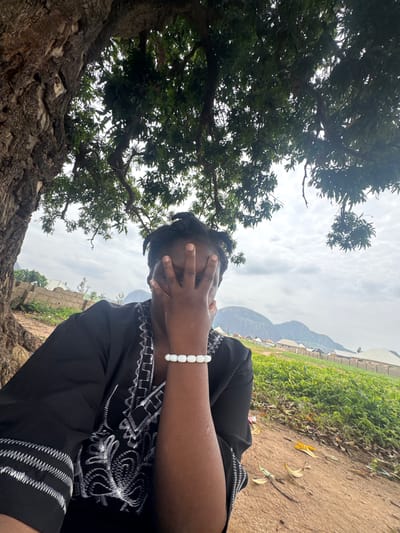 A person sits under a large tree, obscuring their face with their hand. They are wearing a black garment with white embroidered details and a white beaded bracelet on their left wrist. The background shows a rural landscape with buildings, green vegetation, and hills in the distance. The scene appears to be in Maraba, Nigeria, given the presence of these distinctive geological formations and the style of the buildings. The sky is cloudy.