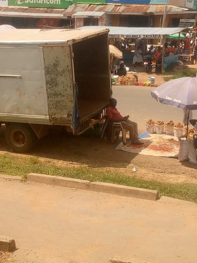 The image depicts an outdoor market scene in South Kabras ward, Kenya, under daylight conditions.
In the foreground, a light-colored, weathered box truck with an open rear door is parked on a dirt shoulder adjacent to a light-brown asphalt road and a concrete curb. A blue cloth hangs from the truck's rear frame. To the immediate right of the truck, a man wearing a red collared shirt and dark trousers is seated on a white plastic stool, looking down at an object in his hands, possibly a mobile phone. Two additional white plastic stools are stacked next to him.
Adjacent to the seated man, a market stall is set up on the dirt ground, partially shaded by a blue umbrella displaying a bank logo, which includes the word "Bank." The stall features several white plastic buckets filled with light-brown, round produce, along with numerous small, red, elongated items (likely chili peppers) arranged on a white tarp. Two partially visible white sacks are also present.
In the mid-ground, across the asphalt road, a bustling market area is visible. This area contains multiple structures with corrugated metal roofs, some exhibiting rust. One structure has a visible sign stating "DONATED BY MULAMULA ZACHARIA." Several individuals are seen standing and seated within this market area. A green "Safaricom" sign is partially visible at the top left, and another small sign possibly indicating "PORK" is present. The ground throughout the market area is unpaved, consisting of dirt. Shadows suggest direct sunlight.