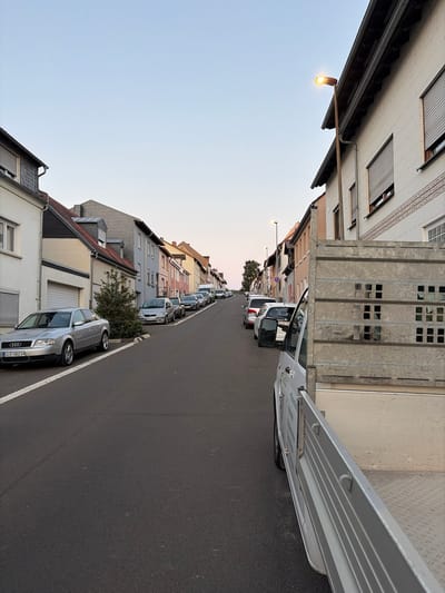 The image shows a street scene in Kirchheimbolanden, Germany, during the late afternoon or early evening. The sky is clear and a pale blue. The street is paved and has a white line marking the parking area on the left side.
On the left side of the street, a silver Audi sedan is parked, facing away from the camera. Further down the street, a row of parked cars is visible. The buildings lining the street are a mix of residential and commercial, with some featuring white facades and dark roofs. One building has a prominent street lamp extending from its side, which is illuminated.
On the right side of the street, a silver utility truck with an open, metal-sided cargo bed is parked. The cargo bed has a metallic grid pattern with square openings. The truck is facing towards the camera, but its side is the primary focus in the foreground. Some text is partially visible on the side of the truck.
The overall impression is a quiet residential street, possibly in a suburban area, with cars parked along the road and buildings on either side. The lighting suggests the transition from day to evening.