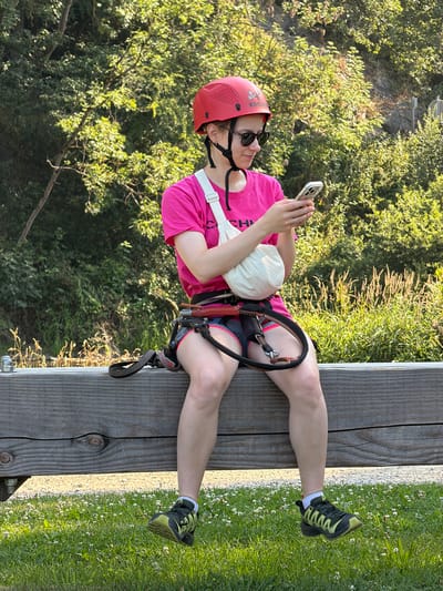 A person, wearing a pink t-shirt, red helmet, sunglasses, and a harness, sits on a wooden platform. They are holding a smartphone in their hands and looking down at it. The person's legs are bent, and they are wearing athletic shoes.
The background consists of lush green trees and foliage, suggesting an outdoor setting, possibly a park or a natural reserve. The sun appears to be shining, casting light and shadows, indicating it is daytime. The overall scene appears to be calm and relaxed, with the person engrossed in their phone.
Text visible on the helmet reads "EDELRID". The person's t-shirt has some text on it, but it is partially obscured and difficult to read. There are also some wildflowers growing in the grass next to the wooden platform.