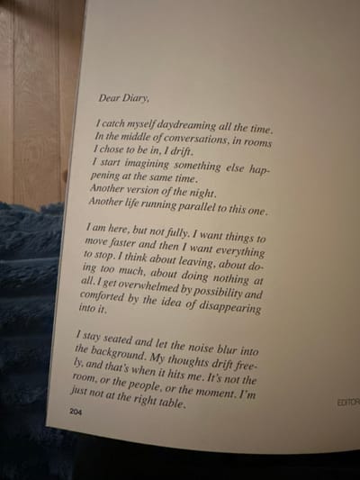 A light-colored open book, displaying page 204, is centrally positioned in what is stated to be a location within the city of Les Lilas, France. The page features printed English text, beginning with "Dear Diary," and details introspective thoughts about daydreaming, parallel lives, feelings of being overwhelmed, and social displacement. A partial word, "EDITOR", is visible at the bottom right edge of the page. The book rests on a dark blue, textured fabric in the foreground, with a light brown wooden surface, exhibiting distinct grain and panel lines, visible in the background to the left and top. No people or actions are depicted.
