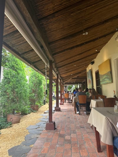 The image depicts a patio dining area located at Traversia 500, Santa Ana, Costa Rica (latitude: 9.934, longitude: -84.179). The patio is covered by a wooden roof with a thatched-style ceiling. The flooring is comprised of red brick tiles. Tables with white linens and chairs are set up on the patio, with people dining. The scene is framed by a wooden support beam. The patio faces a green grassy area lined with potted plants, with a stone pathway that extends along the length of the patio. The background features a whitewashed wall with paintings on display and a glimpse of foliage.