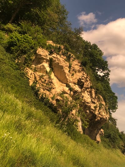 The image shows a rocky outcrop in a natural environment. The outcrop is light tan in color and appears to be composed of fractured stone. Green vegetation, including trees, bushes, and grasses, surrounds the rock formation. The sky is visible in the upper left corner, displaying patches of blue sky interspersed with white clouds. The scene appears to be taken on a hillside. The location is in the city of Bolsover, United Kingdom.