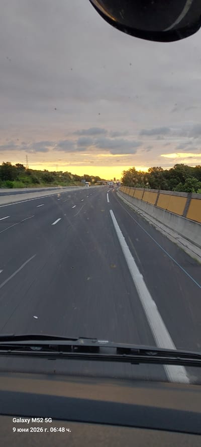 This image captures a view from inside a vehicle driving on a highway at sunset. The sky is filled with dramatic clouds illuminated by the setting sun, casting a warm orange and yellow glow. The highway stretches ahead, with multiple lanes marked by white lines. Trees and greenery line the sides of the road, and a yellow sound barrier is visible on the right. In the distance, several trucks can be seen on the highway. The bottom of the image shows the dashboard and windshield of the vehicle, with a watermark indicating "Galaxy M52 5G" and the date and time "9 юни 2026 г. 06:48 ч." This image was taken on the A9 motorway near Le Boulou, France, a common route for heavy goods vehicles traveling between Spain and Northern Europe.