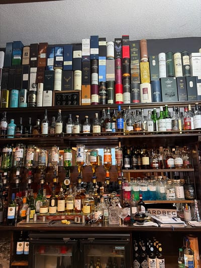 This image provides a detailed view of a well-stocked bar within the Sligachan Hotel, located in Sconser, Isle of Skye, Scotland, United Kingdom. No individuals are present.

The scene features multiple rows of wooden shelving displaying a vast collection of alcoholic beverages. The top two rows are predominantly filled with boxed single malt whiskies from numerous distilleries, including Scapa, Caol Ila, Springbank, Tomatin, Talisker, Dalmore, Deanston, Edradour, Glenfarclas, Glengoyne, Glenfiddich, Highland Park, Jura, Lagavulin, Laphroaig, Macallan, Oban, Old Pulteney, and Glenmorangie. A small wooden plaque labeled "BEDROOMS" with eight circular numbered indicators (Nº1-Nº8) is interspersed among these boxes.

Below the boxed whiskies, two open shelves display a wide selection of bottled spirits, mainly whiskies and some gins. Brands visible include Glen Scotia, Glenlivet, Jura, Laphroaig, Lagavulin, Deanston, Edradour, Cragganmore, Dalmore, Old Pulteney, Talisker, Arran, and Glenmorangie. Several bottles are inverted onto spirit dispensers positioned along the lower shelving.

The main bar counter features multiple operational spirit dispensers containing various liquids, some clear, others colored blue or pink. A dedicated area for cocktail preparation includes metal jiggers, a small cutting board with lemon wedges, and a stirring utensil. A prominent display of gin bottles includes "Misty Isle Gin" (in distinct blue and pink varieties), Harris Gin, Tommy's Gin, Raasay Gin, and Skye Gin. A white sign with "ARDBEG" printed in red is visible on the counter. Additional counter items include stacks of paper, a pen holder with pens, striped straws, and various glassware, including a large snifter.

Beneath the bar counter, two black "POLAR Refrigeration" mini-fridges are visible. A small collection of "Santa Helena" wine bottles and bottles of "Skye Black" and "Thistly Cross" cider/beer are stored in this lower section. The wall behind the shelving features a light, textured surface.