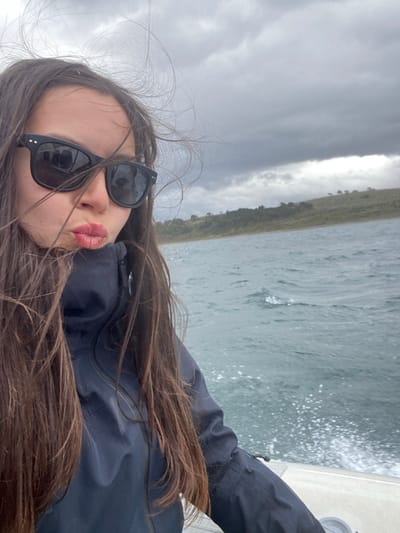 A young woman with long brown hair and sunglasses is on a boat. She wears a dark jacket and poses with her lips puckered. Behind her, water churns from the boat's movement, and a distant shoreline with vegetation is visible. The sky is overcast with dark, heavy clouds, suggesting a potentially stormy day.