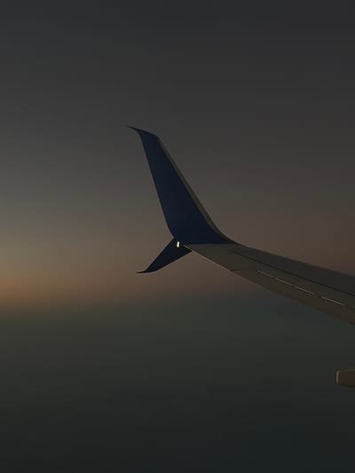 The image shows a portion of an airplane wing, likely viewed from inside the aircraft. The wing has a dark grey or black color and features a triangular-shaped winglet at the end. The background is a gradient of grey, with a faint light streak in the lower portion of the image. The scene evokes a sense of flight, but the specific location in the city of Unknown, United States of America, is not identifiable from the image alone.