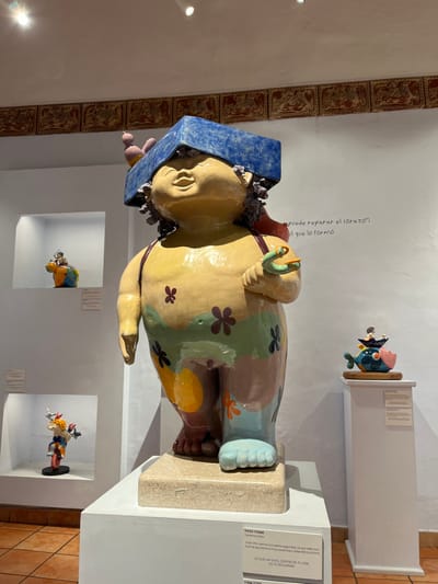 Inside a gallery, a colorful ceramic sculpture of a stout figure stands on a pedestal. The figure wears a blue hat and a flower-patterned garment. Inset wall niches display smaller ceramic pieces. Above the sculpture, handwritten text reads, "Puedo reparar el corazón al que lo formó," which translates to "I can repair the heart to the one who formed it." A nearby sign describes the sculpture as "PASO FIRME," stating "Lo que hay en el centro de tu vida es tu seguridad" or "What is at the center of your life is your security."