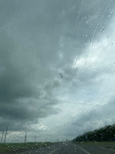 The image depicts a view through a rain-streaked windshield, suggesting travel by vehicle. The sky is overcast with dramatic, dark clouds, indicating impending or ongoing inclement weather. In the distance, a road curves to the right, flanked by green foliage on the right side. On the left, several utility poles are visible in the field. The presence of raindrops on the glass distorts the view of the landscape and sky, creating streaks and blurring details. The scene appears to be set in a rural or semi-rural area, given the open fields and utility poles. No people or specific landmarks of Ilteryakovo, Russia, are identifiable.