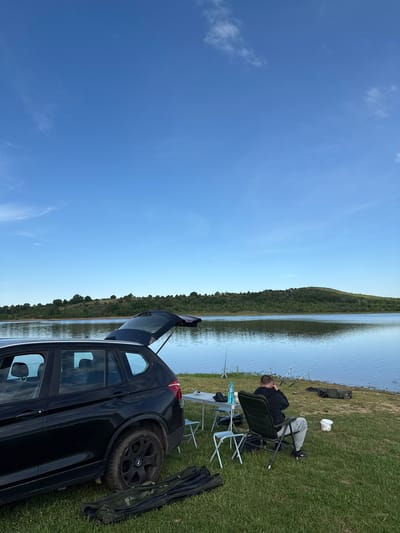 The image shows a man fishing at a lake near Brezovo, Bulgaria. The man is sitting in a chair near fishing rods, a table, and a black BMW. The car's trunk is open. The landscape includes a grassy bank, a body of water, and trees across the water. The sky is blue.