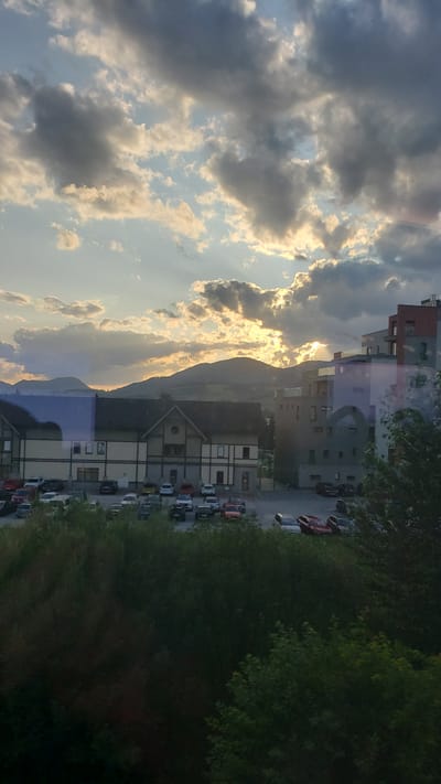 The image depicts a view of the city of Lisková, Slovakia, during sunset. The sky is filled with dramatic clouds, illuminated by the setting sun peeking through. In the foreground, lush green trees partially obscure a view of a parking lot filled with numerous cars. Behind the parking lot, there are several buildings, including a multi-story modern apartment complex with a red-toned facade and a more traditional building with a dark roof and light-colored walls. In the distance, rolling hills and mountains form the backdrop, their outlines softened by the twilight. The overall scene captures a serene, yet dynamic, natural and urban landscape at the end of the day.