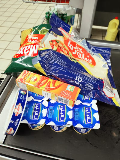 The image displays a collection of grocery items placed on a black surface, possibly a conveyor belt at a supermarket checkout. The items are diverse, including packaged flatbreads, a box of harissa, several yogurt cups, and a bag of chips.
In the foreground, there are eight small yogurt cups with blue and white packaging, labeled "Naty" and "Yaourt." Next to them is a rectangular package of "President" portions, also with blue and white branding, indicating "8 Portions." Behind these, a bright yellow box with red chili peppers depicted on it is visible, labeled "SICAM HARISSA du Cap Bon."
Further back, a large blue and yellow plastic package contains flatbreads, with Arabic and French text. Some of this text indicates "10 Pièces" and "Poids Net: 600g." To the left, a green bag with a red and white label is partially visible, with text that appears to say "Max Chips" and "SPECIAL."
The overall scene suggests a shopping trip in a grocery store. The lighting is bright, typical of indoor retail environments, and there are no indicators of the time of day or weather. The setting is clean and organized, with the tiled floor of a supermarket visible in the upper left corner. The items are neatly arranged, suggesting they are about to be scanned or have just been placed on the conveyor. No people are visible in the image.