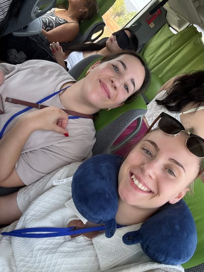 In this selfie-style photo, two young women are seated next to each other on what appears to be a bus or tour vehicle. The woman in the foreground on the right is smiling broadly, her head tilted slightly and supported by a blue travel pillow. She is wearing sunglasses pushed up on her head and a white textured shirt. A blue lanyard is visible around her neck. To her left, another woman is also smiling, looking towards the camera. She is wearing a light beige t-shirt and has a red nail polish on her fingernails. A blue lanyard is also around her neck.

In the background, a third person is visible wearing a black eye mask. Further in the background, another woman with glasses and a patterned top is seated. The setting appears to be daytime, likely warm given the clothing choices. The green curtains of the vehicle are visible, and a hint of greenery and sunlight outside suggests an outdoor journey. The overall mood is cheerful and relaxed, indicating a group enjoying a trip or outing in Albufeira, Portugal. No discernible text is visible in the image.