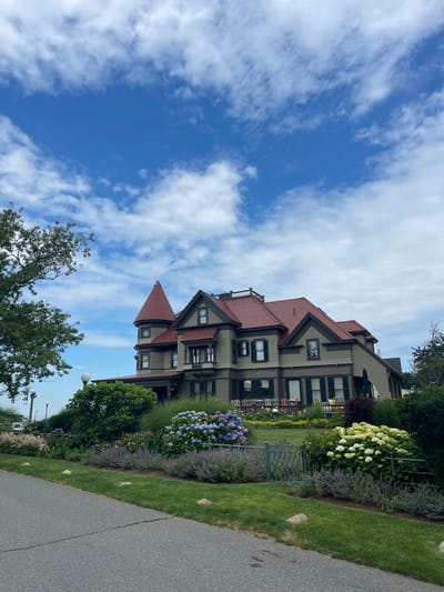 The image depicts a large, historic, Victorian-style house in Oak Bluffs, Massachusetts, USA. The house has a prominent red roof, pale green siding, and dark green shutters. A distinctive conical tower with a red roof is visible on the left side of the house. The property is surrounded by lush landscaping, including vibrant hydrangeas in shades of blue and white, lavender-colored flowers, and tall green grasses. A paved pathway runs along the front of the property, with a decorative metal fence separating the garden from the pathway. The sky above is a clear blue with scattered white, fluffy clouds. A large tree with green leaves is visible on the left side of the frame, obscuring part of the sky. The overall scene suggests a well-maintained estate on a pleasant day.