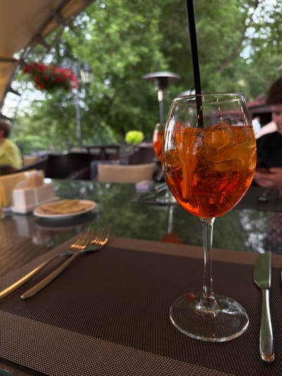 An orange cocktail with ice and a slice of orange sits in a wine glass with a black straw, placed on a table in an outdoor cafe. In the background, out of focus, are trees, other tables, chairs, and people. The scene appears to be set in a restaurant in Izmailovo, Russia, given the presence of a heater which is common in outdoor dining areas during cooler seasons. A fork and knife are laid out on a placemat to the left of the cocktail.