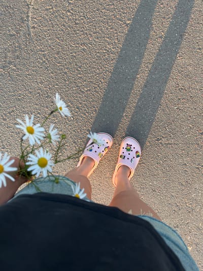 This media file features a close-up, overhead view of a person's legs and feet standing on a textured asphalt surface. The person is wearing light pink Crocs adorned with colorful Jibbitz, including cartoon characters and animals. They are also wearing denim shorts with frayed edges and a dark garment, possibly a skirt or dress, partially obscuring their legs.
In the foreground, a small bouquet of daisies is held by the person, their white petals and yellow centers a stark contrast to the grey asphalt. The lighting suggests it is late afternoon or early evening, with long shadows cast diagonally across the ground from an unseen object. The overall mood is peaceful and casual, hinting at a relaxed outdoor activity. The location context provided is "Пушкино, Russia," which may indicate where the photo was taken. No visible text is present in the image.