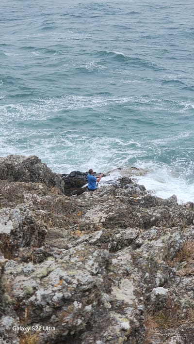 A person is fishing from a rocky outcrop near the sea. The person is wearing a blue t-shirt and sunglasses and is holding a fishing rod. The sea is choppy with waves breaking on the rocks. The foreground is dominated by large, moss-covered rocks. This scene is located in Cemaes, United Kingdom.