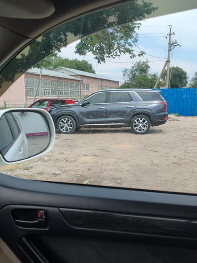 A dark gray SUV is parked on a dirt lot next to a light pink building with large, multi-paned windows. A red sedan is parked in front of the building. A bright blue fence is visible behind the SUV. The scene appears to be in an industrial or commercial area, possibly in Novopavlovka, Kyrgyzstan, given the presence of utility poles and a somewhat rough terrain. The image is taken from inside another vehicle, with a side mirror reflecting part of the interior. The foreground shows the black interior of the car, including a door handle with a red accent. The sky is overcast, and trees are visible in the background.