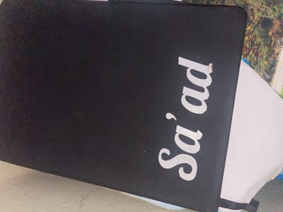 The image shows a black laptop with white text that reads "Sa'ad" on its lid. The laptop is closed and positioned at an angle. The background is blurred, but appears to be a mix of natural elements, possibly foliage, and a white surface. The scene is indoors or on a surface that is not directly exposed to the elements. There are no people visible in the image. The lighting suggests it is daytime, with the light source coming from the side, creating shadows on the laptop lid. The overall scene is casual and appears to be a close-up of an object. The location in Safari, Nigeria, is not discernible from the visual information provided in the image.