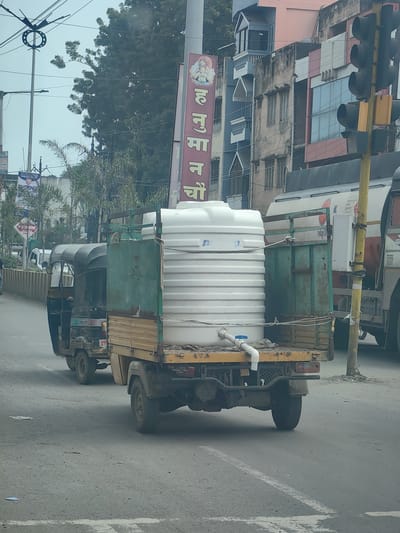 The image captures a street scene in Datala, India, featuring a three-wheeled vehicle laden with a large white water tank. The vehicle, a common mode of transport in the region, has a rustic, yellow and green painted body and is parked on an asphalt road. In the background, buildings of varying architectural styles and heights are visible, some with pastel-colored facades and balconies. A prominent red and white sign with Hindi script, likely advertising a business or service, stands vertically. Traffic signals and utility poles are also present, indicating a developed urban setting. The sky is overcast, suggesting a cloudy day. No people are clearly discernible, and the focus is on the vehicle and its cargo, along with the surrounding urban environment.