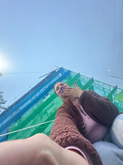 A woman with bright blue nail polish is blowing a kiss at the camera in front of a building undergoing renovation, draped in blue and green scaffolding nets. She is wearing a brown fuzzy jacket over a pink top. The sky is a clear, bright blue. The scene appears to be outdoors in an urban environment.