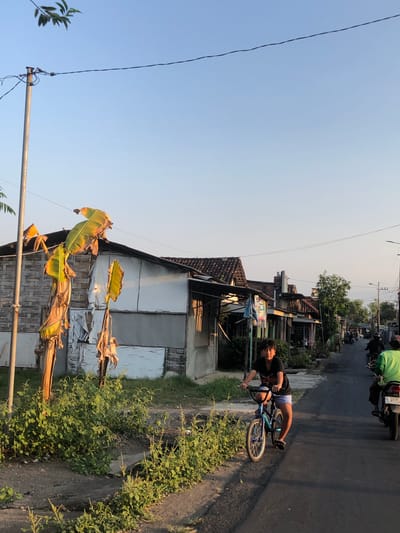 The image depicts a street scene in the city of Glagah Arum, Indonesia. A young person is riding a BMX bicycle down the paved street, wearing a black t-shirt and blue shorts. The street is lined with single-story buildings, some of which appear to be shops or residences. Banana trees with large, yellowing leaves are visible to the left of the street, near a utility pole. In the background, several motorcycles are also present on the street, with riders on them. The sky is clear and blue, suggesting it is daytime, likely late afternoon given the warm lighting.