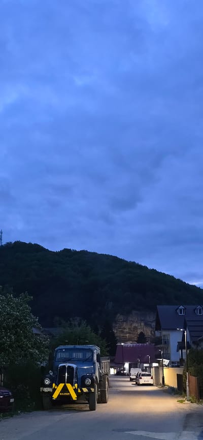 A vertical shot depicts a street scene in Kolašin, Montenegro at dusk or night. The sky, which fills much of the left side of the frame, is a uniform dark blue with faint cloud cover. A densely wooded hillside occupies the upper-center of the image. Buildings, including houses with dormer windows, are situated along the right side of the street. Several cars and a van are parked on the street. The foreground is dominated by a blue Saurer truck, angled towards the viewer. It bears yellow and black hazard stripes on its front bumper.