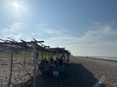 The image depicts a beach scene at Casitas, Mexico, under a bright, sunny sky with scattered clouds. In the foreground, a group of people are seated at a table under a makeshift thatched-roof shelter on a sandy beach. The shelter is supported by wooden poles. Several individuals are visible sitting on chairs around the table, with drinks and a cooler. Further down the beach, other beachgoers can be seen, some near umbrellas and the ocean. The sea has moderate waves, and the shoreline stretches into the distance. Vegetation is present behind the shelter, and the sand is dark.