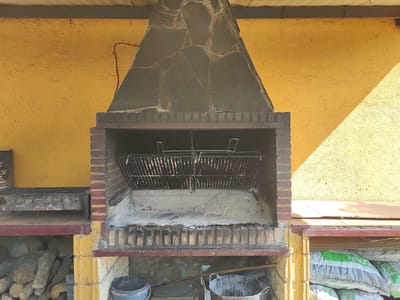 The image shows an outdoor brick barbecue grill against a textured yellow wall. The grill has a stone facade and a metal grate. To the left of the grill, there is a wooden shelf holding firewood, and to the right, there are several bags of what appears to be soil or compost. In the foreground, two buckets are visible, one grey and one metallic. The scene appears to be located in Cadalso, Spain, as indicated by the context provided. There are no people or animals present in the image. The lighting suggests it is daytime, with sunlight casting shadows on the yellow wall.
