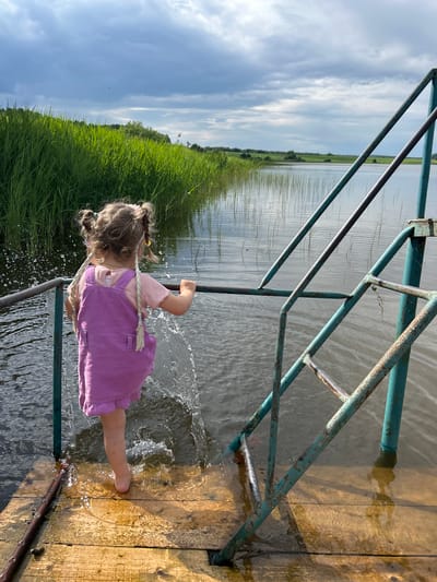 A young child with braided pigtails, wearing a purple dress and pink shirt, stands on a wooden platform at the edge of a body of water. The child is splashing water with their foot. A metal railing and ladder, painted teal and showing signs of rust, are positioned to the right of the child. The background features a dense area of tall green grass bordering the water, with a distant landscape of trees and a cloudy sky. The scene appears to be outdoors, likely in a natural setting near a river or lake, consistent with the rural environment of Верх-Рождество, Russia.