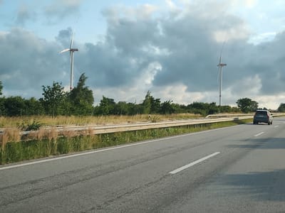 The image captures a view from a moving vehicle on a highway. In the foreground, the asphalt road with white lane markings dominates the lower portion of the frame. To the left of the road, a metal guardrail separates the highway from a grassy embankment dotted with tall, dry grasses and green vegetation. Beyond the embankment, a line of trees forms a dense backdrop. Two wind turbines are visible in the distance, one on the left and another further to the right, both silhouetted against a cloudy sky. A dark-colored SUV is seen driving ahead on the right lane of the highway. The scene suggests a rural or semi-rural setting, likely in the vicinity of Gremersdorf, Germany, given the presence of wind turbines and highway infrastructure. The lighting indicates it is daytime, possibly late afternoon, with overcast conditions.