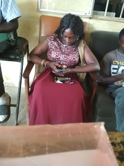 The image shows three people seated indoors. In the foreground, a woman with dark curly hair and wearing a maroon and white patterned top and a maroon dress is seated in a chair. She is holding a smartphone in her right hand and looking down at it. To her left, a person wearing a light blue and white striped shirt and dark pants is seated on a stool. To the woman's right, a person wearing a dark grey t-shirt with yellow lettering and blue jeans is seated in a chair. The environment appears to be a waiting area, possibly in a government building or clinic, with simple chairs and a table in the foreground. The walls are painted a light yellow. The location is identified as Bisichi, Nigeria.