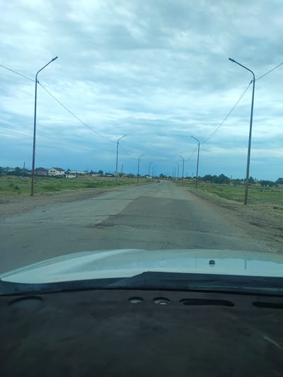 The image shows a view from inside a vehicle looking out at a road lined with streetlights and power lines in Ayagöz, Kazakhstan. The road appears to be asphalt and is in a state of disrepair with visible cracks and potholes. To the left and right of the road, there is dry, grassy terrain. In the distance, on the left side of the road, there are several houses and other buildings that appear to be part of a residential area. On the right side, there are some structures that resemble ruins or unfinished buildings. The sky is overcast with clouds. A single vehicle is visible on the road in the distance. The dashboard and windshield of the vehicle from which the photo is taken are visible in the foreground.